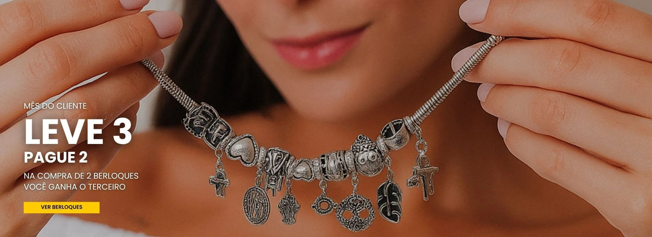 Banner principal com promoção Leve 3 Pague 2 de berloques. Mulher segura uma pulseira de charms de prata cheia de pingentes.